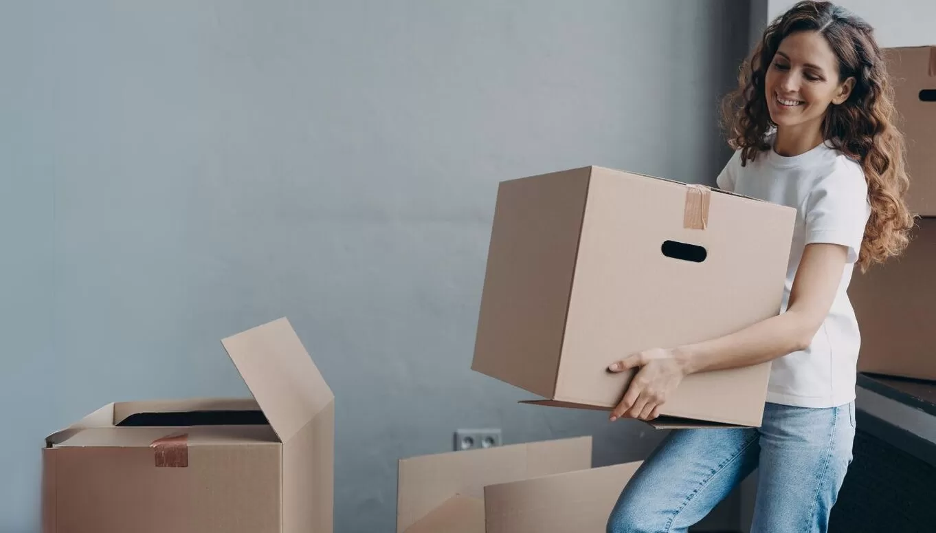 A smiling woman in casual clothing lifting a cardboard box in a room with multiple boxes, suggesting moving or organizing.